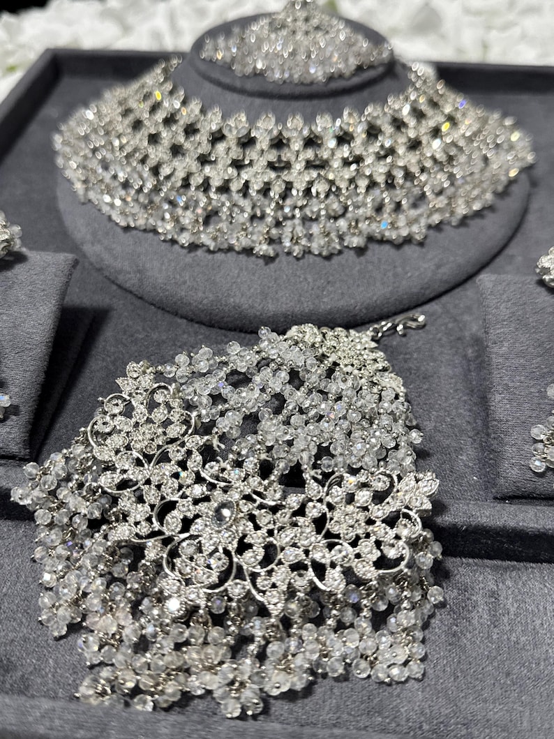 Puede incluir: Un deslumbrante conjunto de joyas de novia con tiara, collar y pulsera. El conjunto est&aacute; hecho de cristales transparentes y brillantes dispuestos en un intrincado dise&ntilde;o floral. Las joyas se exhiben sobre una superficie de terciopelo gris.