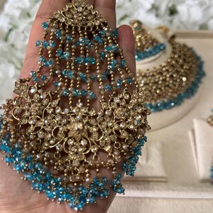 Puede incluir: Un conjunto de joyas dorado ornamentado, que incluye una pieza para la cabeza con cuentas azules colgantes. El conjunto incluye un collar y pendientes, todos adornados con detalles brillantes. El intrincado dise&ntilde;o muestra una mezcla de estilos cl&aacute;sicos y contempor&aacute;neos, ideal para ocasiones especiales.