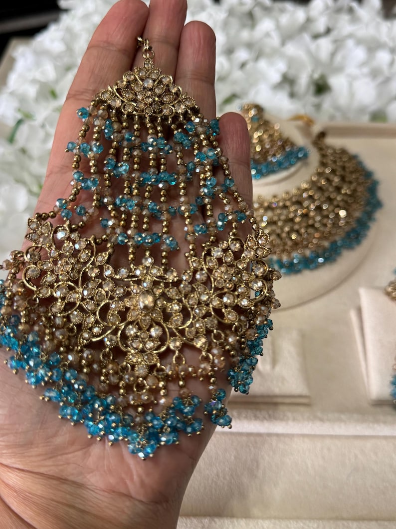 Puede incluir: Un conjunto de joyas dorado ornamentado, que incluye una pieza para la cabeza con cuentas azules colgantes. El conjunto incluye un collar y pendientes, todos adornados con detalles brillantes. El intrincado dise&ntilde;o muestra una mezcla de estilos cl&aacute;sicos y contempor&aacute;neos, ideal para ocasiones especiales.