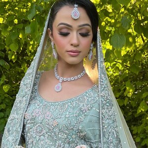 May include: A woman wearing a mint green embroidered wedding dress with a matching veil. She is adorned with a diamond necklace, earrings, and a headpiece, all featuring pink gemstones. The background is a lush green foliage.