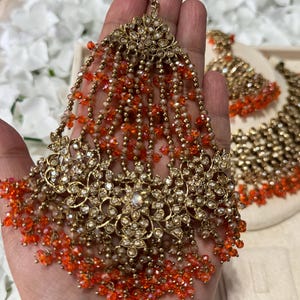 Puede incluir: Un tocado dorado ornamentado con intrincados dise&ntilde;os florales y hebras colgantes de cuentas naranjas y transparentes. La pieza de joyer&iacute;a est&aacute; adornada con peque&ntilde;os cristales brillantes. Un juego de aretes a juego es visible en el fondo.