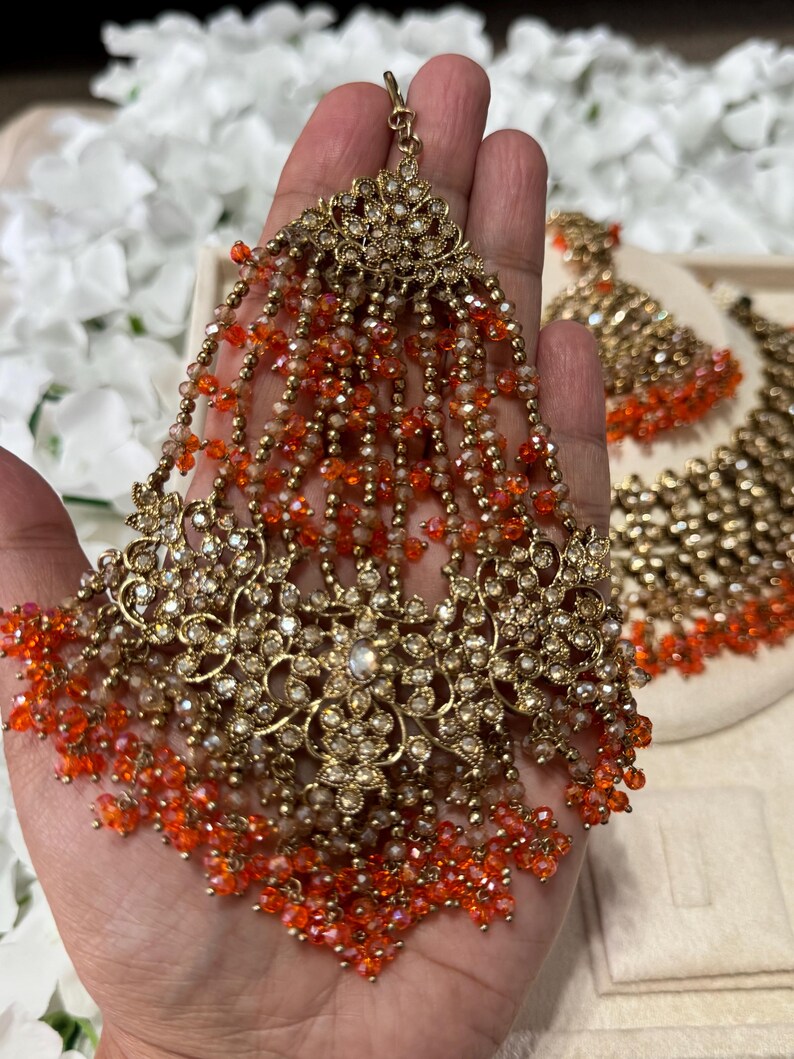 Puede incluir: Un tocado dorado ornamentado con intrincados dise&ntilde;os florales y hebras colgantes de cuentas naranjas y transparentes. La pieza de joyer&iacute;a est&aacute; adornada con peque&ntilde;os cristales brillantes. Un juego de aretes a juego es visible en el fondo.