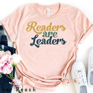 Könnte beinhalten: Ein pfirsichfarbenes T-Shirt mit dem Text "Readers are Leaders" in einer Retro-Schrift. Der Text ist in Gold-, Grün- und Blautönen gehalten.