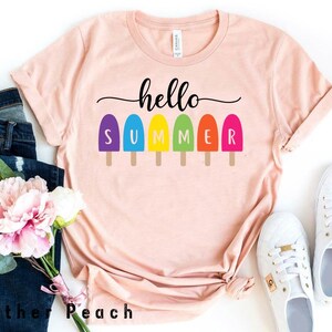May include: A light peach-colored t-shirt with the words "hello SUMMER" in black and colorful popsicle graphics. The shirt is styled with blue jeans, white sneakers, and a bouquet of pink and white flowers.