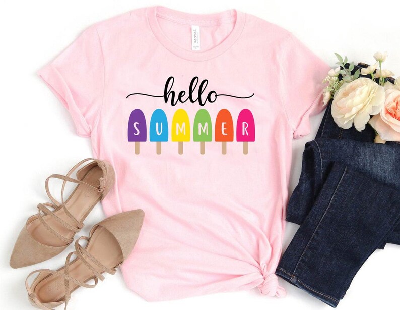 May include: A light pink t-shirt with the words "hello SUMMER" in black script and colorful popsicle graphics. The shirt is styled with a pair of beige flats and dark blue jeans.
