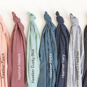May include: A collection of nine folded t-shirts in various colors, including white, peach, mauve, dusty blue, slate, navy, athletic heather, dark grey, and black. Each shirt has a label with its color name.