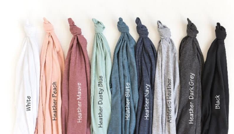 May include: A collection of nine folded t-shirts in various colors, including white, peach, mauve, dusty blue, slate, navy, athletic heather, dark grey, and black. Each shirt has a label with its color name.