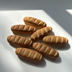 May include: A collection of seven small, brown, oblong-shaped bread loaf miniatures. Each piece features a smooth, rounded surface with four distinct, parallel diagonal score marks across the top, mimicking the appearance of baked artisan bread.