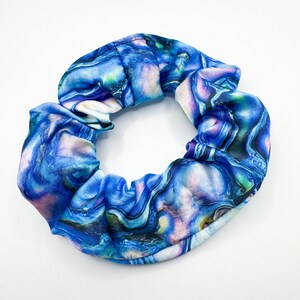 Puede incluir: Una goma para el pelo azul con un patrón arremolinado y veteado en tonos de azul, morado y blanco. La tela está fruncida y elástica, creando una forma circular. Este accesorio para el cabello es adecuado para varios peinados.