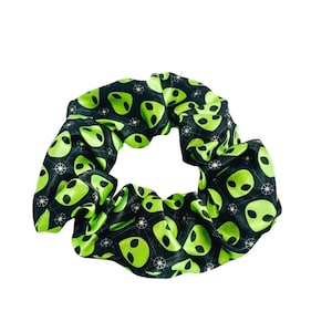 May include: A scrunchie with a black background and a repeating pattern of green alien faces. The scrunchie is made of a soft, gathered fabric, perfect for securing hair in a ponytail or bun. The alien faces are a bright, neon green color.