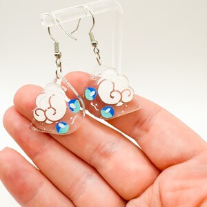 Puede incluir: Un par de pendientes transparentes en forma de nube de acrílico con detalles blancos y azules. Los pendientes tienen ganchos para las orejas de color plateado. El diseño de la nube presenta remolinos blancos y detalles de gotas de lluvia azules y turquesas.