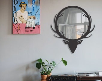 Op de afbeelding: Een zwarte hertenkop-silhouet wandspiegel met een ronde spiegel in het midden. De spiegel reflecteert een kamer met een raam. De spiegel hangt aan een grijze muur boven een wit rek met een platenspeler en een boekenkast.