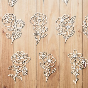 May include: A selection of white, laser-cut floral designs, including roses and other flower types, arranged on a wooden surface. The delicate outlines of the flowers create a minimalist aesthetic, suitable for crafting or decorative purposes.