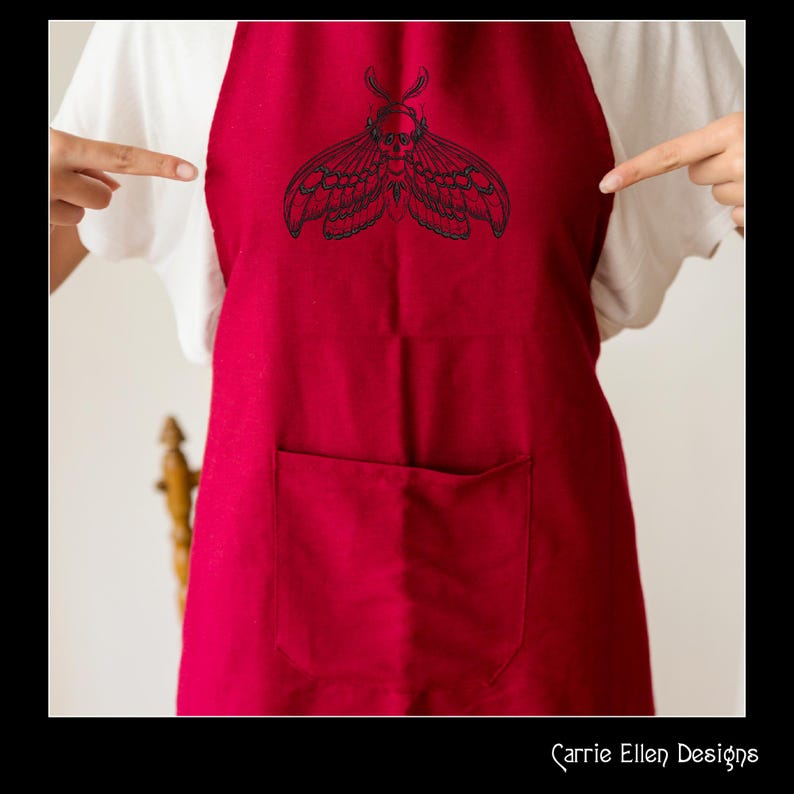 May include: A red apron with a large black moth design embroidered on the front. The apron has a large pocket and is being pointed at by two hands. The text "Carrie Ellen Designs" is at the bottom.