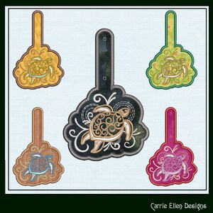 May include: Five different coloured fabric keychains with a decorative embroidered sea turtle design. Each keychain has a loop at the top for attaching keys. The keychains are in yellow, green, black, brown, and pink.