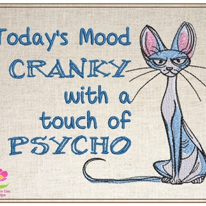 Può includere: Disegno ricamato su uno sfondo di tessuto beige con un gatto blu con orecchie rosa e il testo "Today's Mood Cranky with a touch of Psycho".