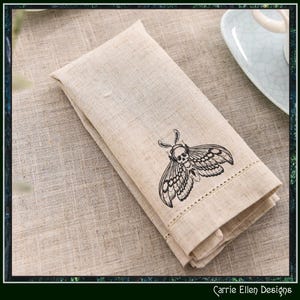 May include: Beige linen napkin with a black moth illustration featuring a skull design. The napkin is folded and has a stitched border. The Carrie Ellen Designs logo is in the bottom right corner.