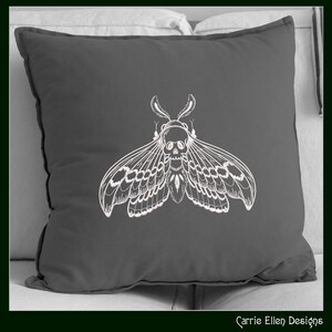 May include: A dark gray square pillow featuring a white, detailed illustration of a moth with a skull-shaped head. The moth's wings are spread, showcasing intricate patterns. The pillow is on a light-colored surface, with the text "Carrie Ellen Designs" in the bottom right corner.