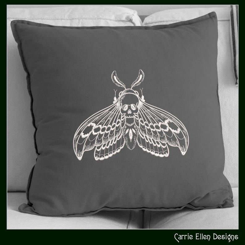 May include: A dark gray square pillow featuring a white, detailed illustration of a moth with a skull-shaped head. The moth's wings are spread, showcasing intricate patterns. The pillow is on a light-colored surface, with the text "Carrie Ellen Designs" in the bottom right corner.