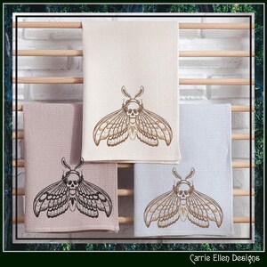 May include: Four decorative kitchen towels in cream, pink, and light blue, each featuring an embroidered moth design with a skull. The towels are displayed on a wooden rack against a white brick wall. The towels are a unique addition to any kitchen.