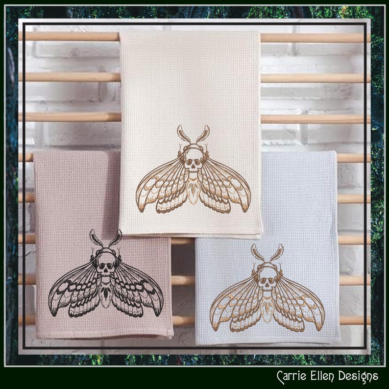 May include: Four decorative kitchen towels in cream, pink, and light blue, each featuring an embroidered moth design with a skull. The towels are displayed on a wooden rack against a white brick wall. The towels are a unique addition to any kitchen.