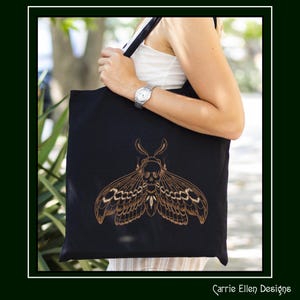 May include: Black tote bag featuring a detailed, gold-colored moth design with a skull motif. The tote bag is held by a person. The words "Carrie Ellen Designs" are printed in the bottom right corner.