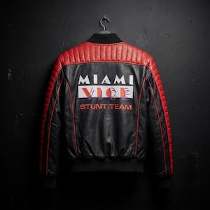 May include: A black and red leather bomber jacket featuring a quilted shoulder design and a ribbed collar, cuffs, and hem. The back displays the white and orange text MIAMI VICE STUNT TEAM printed across the center of the black leather panel.
