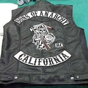 May include: A black leather vest featuring white embroidered patches on the back. The top patch reads SONS OF ANARCHY, the center patch depicts a grim reaper holding a scythe and a rifle, and the bottom patch reads CALIFORNIA with a small MC patch.