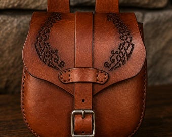 Op de afbeelding: Een bruine leren tas met een voorflap voorzien van in reliëf aangebrachte Keltische knoopmotieven. De tas heeft een verticale leren riem die wordt vastgezet met een metalen gesp en twee leren lussen aan de bovenkant voor bevestiging aan een riem.
