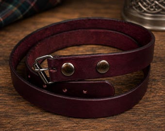May include: A deep burgundy leather belt coiled in a circle. The belt features a smooth finish, a silver-toned metal buckle, and two matching silver-toned metal rivets securing the strap near the buckle. The edges of the leather are dark and defined.