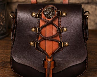 May include: A dark brown leather pouch featuring a lighter tan leather center panel with a decorative lace-up design. The front flap is secured with a leather strap and includes a circular metal emblem with an intricate Celtic knot pattern.