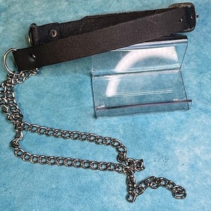 May include: A black leather strap featuring a silver-toned metal D-ring at one end and a matching silver-toned metal chain attached to the ring. The strap includes a metal snap button fastener and a rectangular metal buckle at the opposite end.