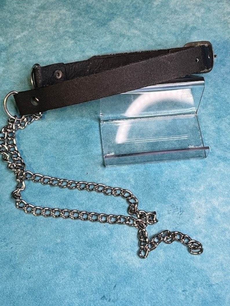 May include: A black leather strap featuring a silver-toned metal D-ring at one end and a matching silver-toned metal chain attached to the ring. The strap includes a metal snap button fastener and a rectangular metal buckle at the opposite end.