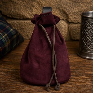 May include: A deep burgundy suede drawstring pouch featuring a soft, matte texture. The bag is cinched at the top with a dark brown leather cord that hangs down in two thin, knotted strands. A black leather loop is attached to the top center.