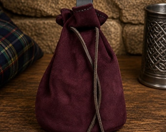 May include: A deep burgundy suede drawstring pouch featuring a soft, matte texture. The bag is cinched at the top with a dark brown leather cord that hangs down in two thin, knotted strands. A black leather loop is attached to the top center.