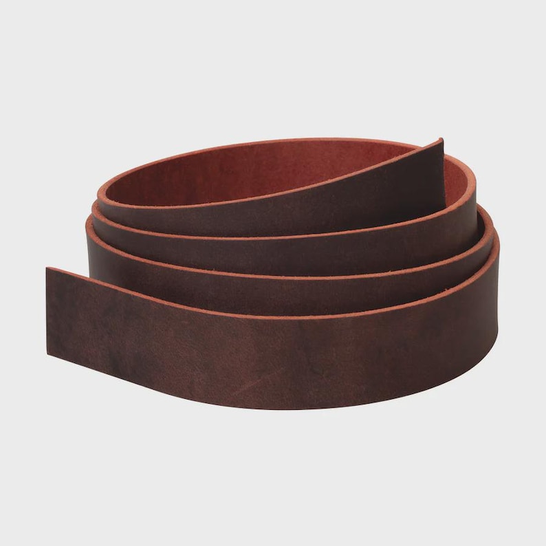 May include: A long, continuous strip of dark brown leather coiled into a circular shape. The leather features a smooth, matte exterior surface with a visible grain and a contrasting reddish-brown interior side, showing the natural texture of the material.