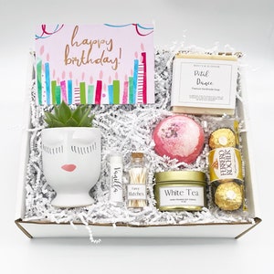 May include: A gift box filled with a succulent in a face-shaped pot, a 'happy birthday' card, a bar of soap, a bath bomb, a lip balm, matches, a candle, and Ferrero Rocher chocolates. The box is filled with white paper shreds.