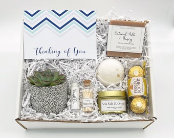 May include: A gift box filled with a succulent plant, a card that says "Thinking of You", a bar of soap, a bath bomb, a candle, lip balm, matches, and Ferrero Rocher chocolates. The card has a blue and white chevron pattern.