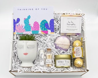 May include: A gift box filled with a variety of items. Includes a succulent in a white face planter, a card that says "Thinking of You", a bar of lavender soap, a bath bomb, a candle, lip balm, matches, and Ferrero Rocher chocolates.