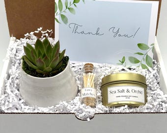 May include: A gift box containing a succulent in a white ceramic pot, a small glass bottle of fancy matches, and a gold-colored candle labeled "Sea Salt & Orchid". A card with "Thank You!" is also included.