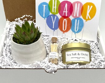 May include: A gift box containing a white ceramic succulent planter, a small glass bottle of matches, a gold-colored candle tin, and a card that says "Thank You" with balloon graphics. The candle is labeled "Sea Salt & Orchid".