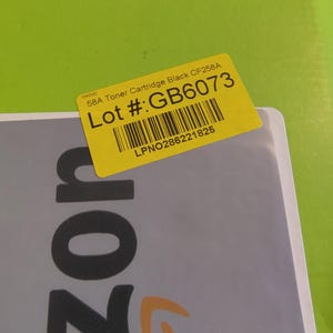 May include: A yellow sticker with the text "58A Toner Cartridge Black CF258A" and "Lot #: GB6073" is affixed to a gray surface. The sticker also includes a barcode and the text "LPNO286221825". The background is a bright green.