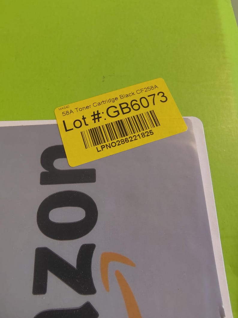 May include: A yellow sticker with the text "58A Toner Cartridge Black CF258A" and "Lot #: GB6073" is affixed to a gray surface. The sticker also includes a barcode and the text "LPNO286221825". The background is a bright green.