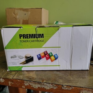 May include: A white and green box labeled "PREMIUM TONER CARTRIDGE" with a diagram of a pie chart, coins, and a pen. The box also has the word "SUCCESS" spelled out with colorful blocks. A brown cardboard box is in the background.