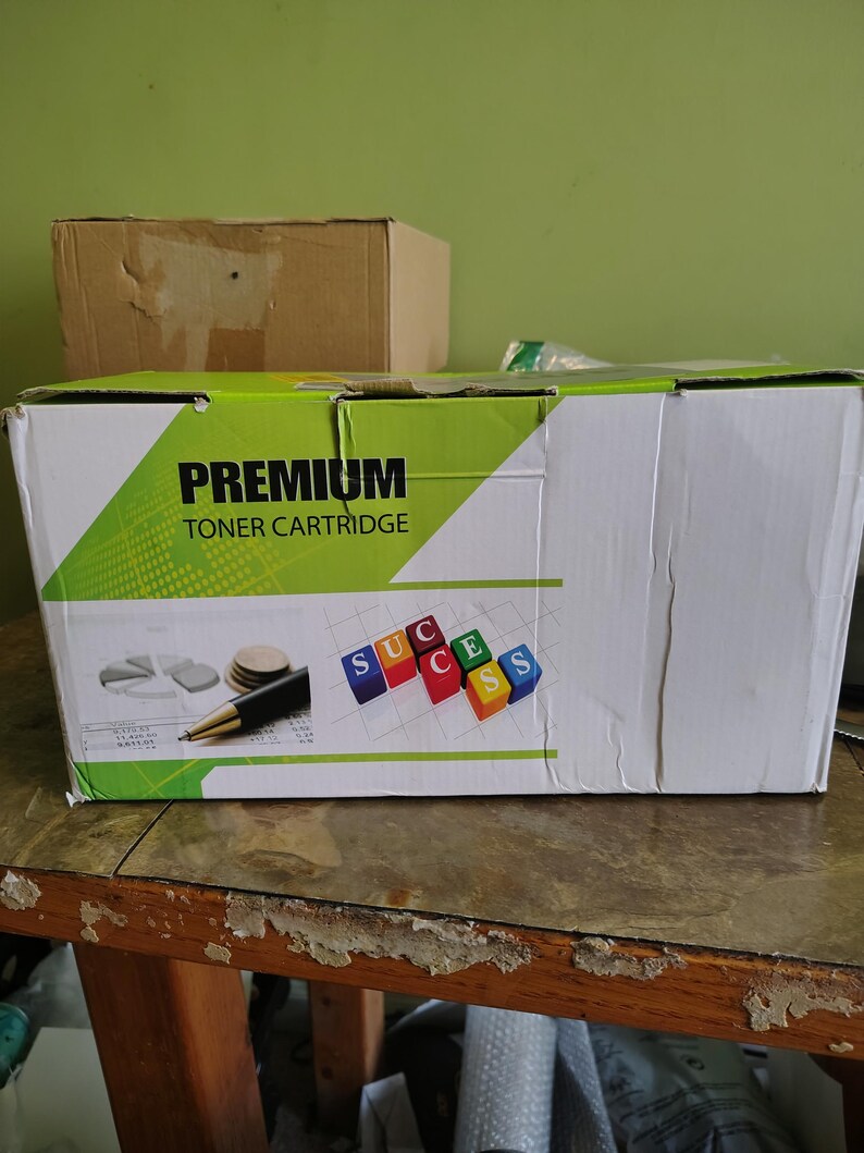 May include: A white and green box labeled "PREMIUM TONER CARTRIDGE" with a diagram of a pie chart, coins, and a pen. The box also has the word "SUCCESS" spelled out with colorful blocks. A brown cardboard box is in the background.