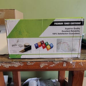 May include: A cardboard box labeled "PREMIUM TONER CARTRIDGE" with green and white accents. The box features the words "Superior Quality," "Excellent Reliability," and "100% Satisfaction Guaranteed." The word "SUCCESS" is spelled out with colorful blocks.
