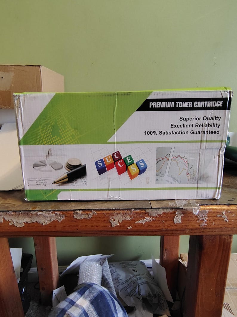 May include: A cardboard box labeled "PREMIUM TONER CARTRIDGE" with green and white accents. The box features the words "Superior Quality," "Excellent Reliability," and "100% Satisfaction Guaranteed." The word "SUCCESS" is spelled out with colorful blocks.