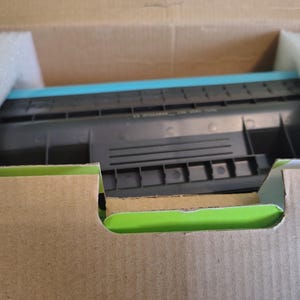 May include: A new printer cartridge, black and teal, is nestled inside a cardboard box with protective foam. The box is brown with a green interior edge. The cartridge is the primary focus.