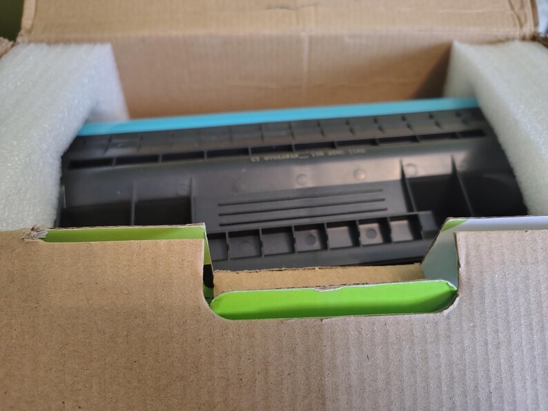 May include: A new printer cartridge, black and teal, is nestled inside a cardboard box with protective foam. The box is brown with a green interior edge. The cartridge is the primary focus.