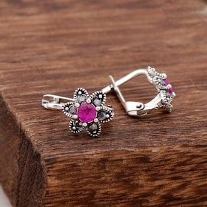 May include: A pair of silver earrings with a pink stone in the center of each earring. The earrings are shaped like a flower with small black stones around the pink stone.